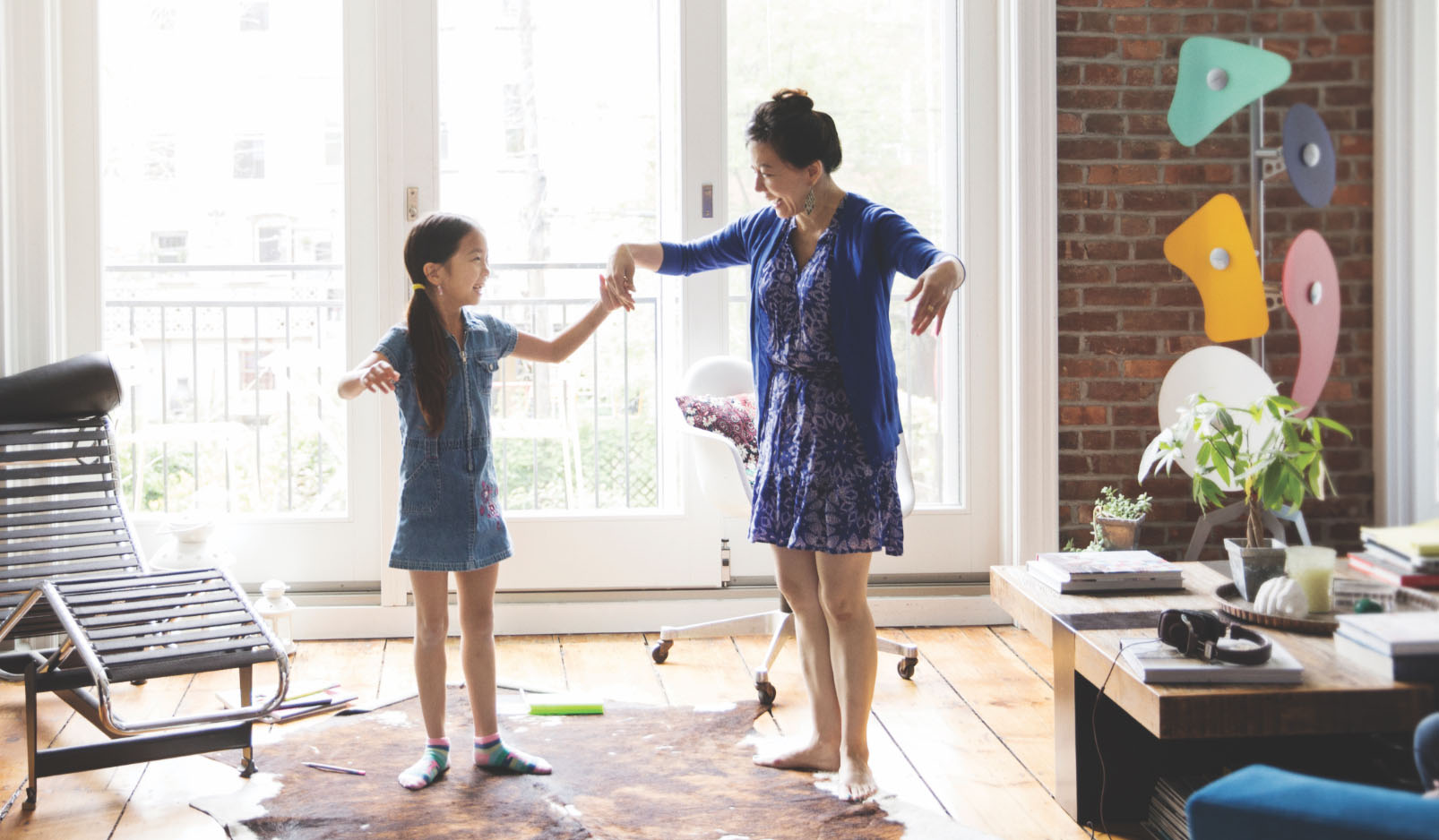 A woman and a young girl dance together in a sunlit living room, smiling and holding hands.