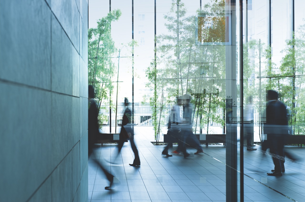 Blurred figures of people walking through a modern building with large windows and green plants in the background.